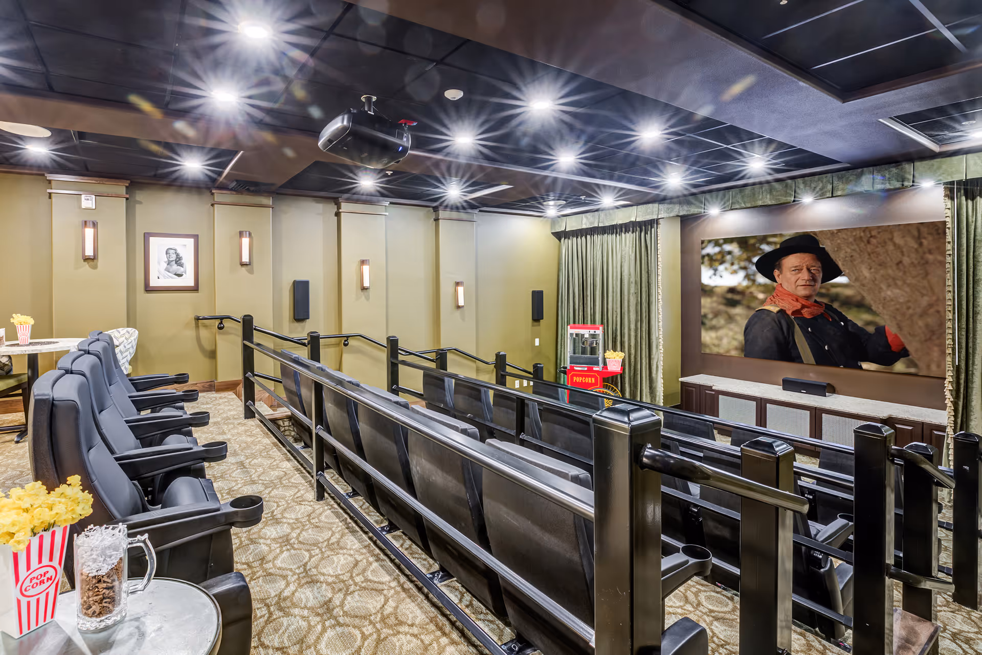Interior view of a small movie theater room with several rows of black cushioned seats, a large screen showing a man in a cowboy hat, a popcorn machine, and popcorn containers on tables. The room has beige walls, carpeted floors, and ceiling lights.