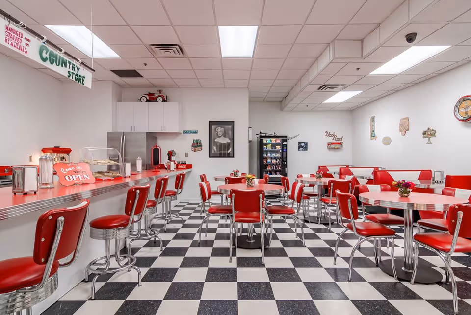 A retro-style dining area with red and chrome chairs and tables, black and white checkered floor, a counter with stools, and vintage decor on the walls. There is a sign that reads 'Country Store' and a small display case with baked goods on the counter.