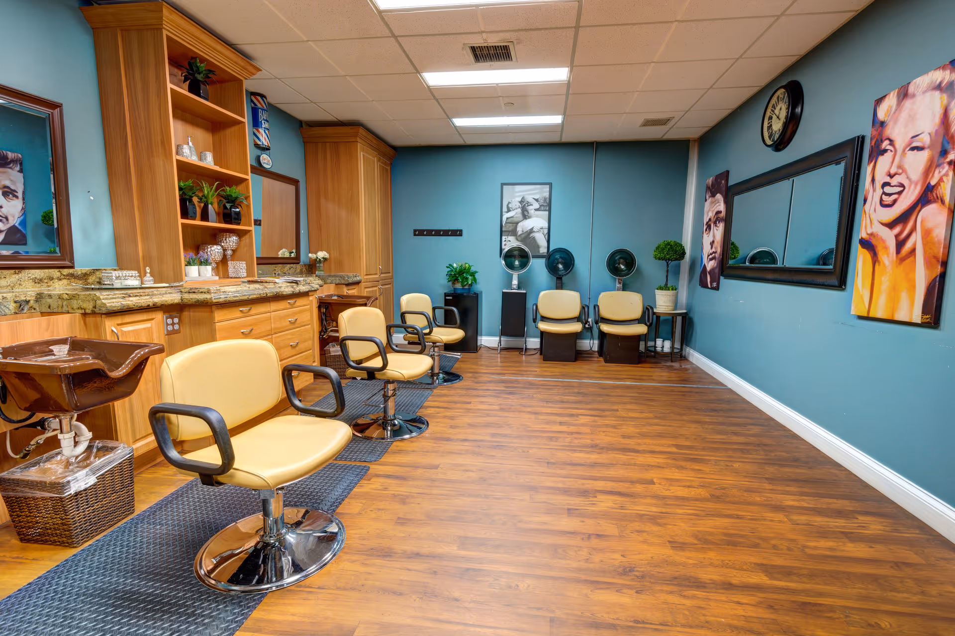 Interior view of a salon area with several beige salon chairs, hair washing station, wooden cabinets with granite countertops, and blue walls decorated with framed portraits and a clock.