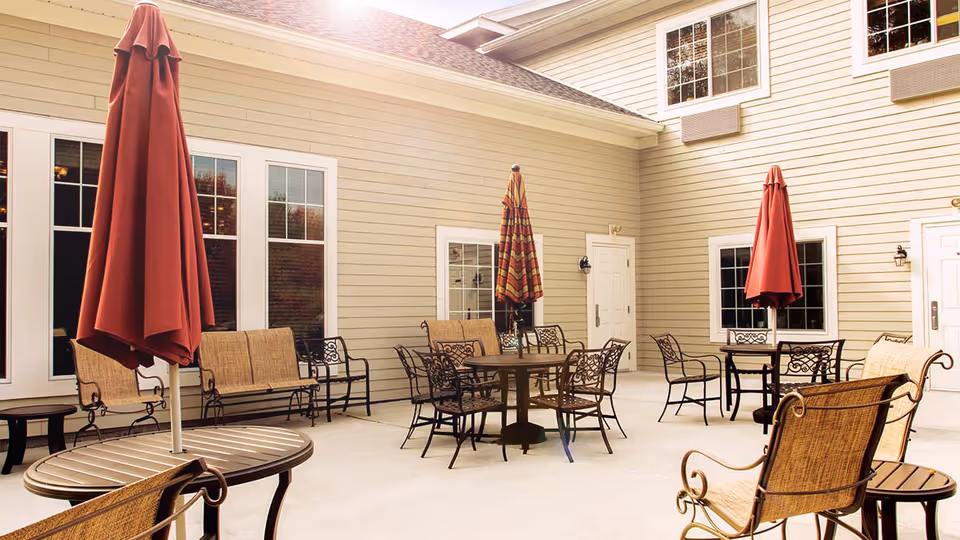 Outdoor patio area with several round tables and chairs, each table having a closed red or striped umbrella. The patio is surrounded by beige siding walls with multiple windows and doors.