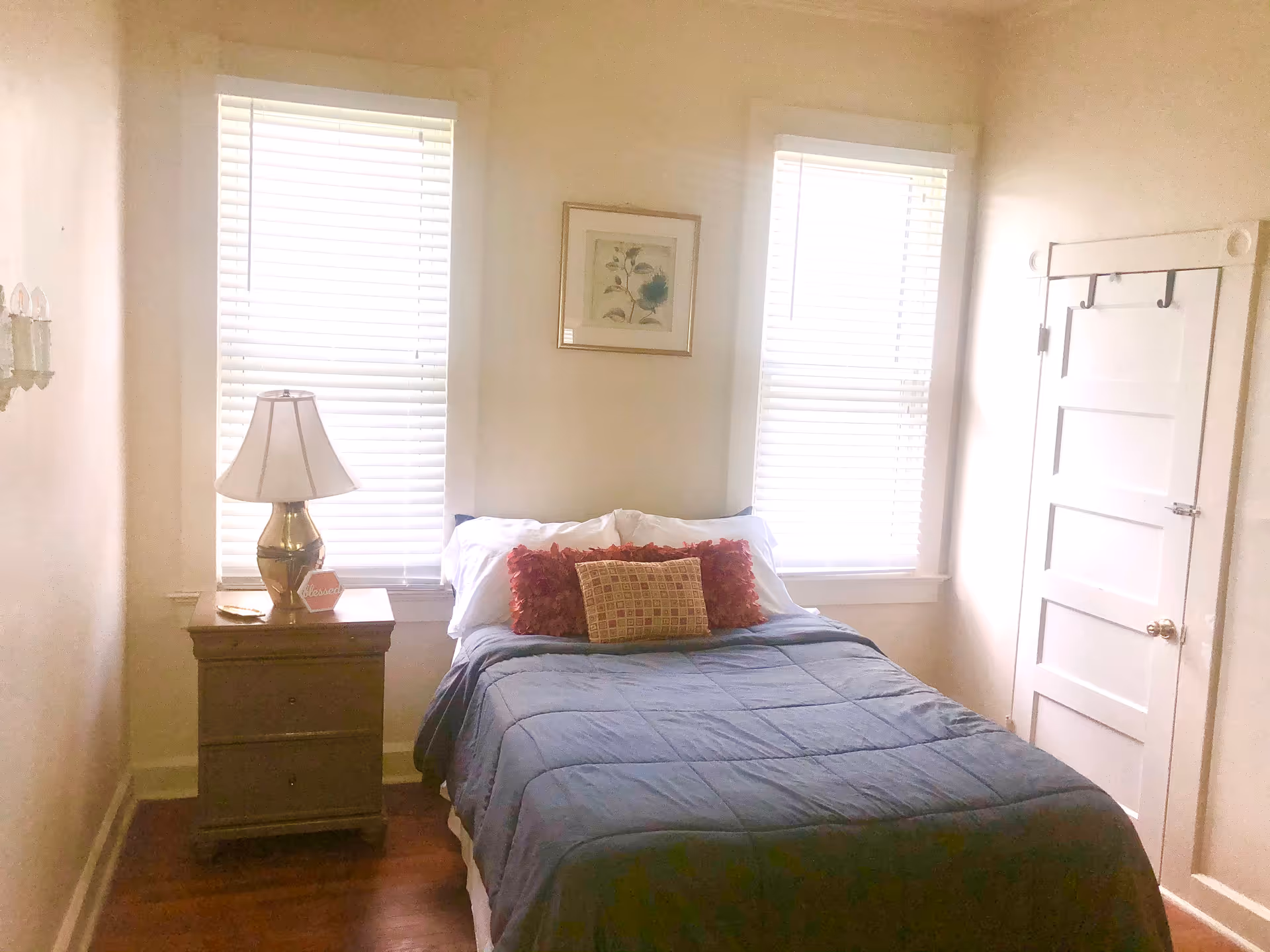 A small bedroom with a neatly made bed featuring a blue comforter and decorative pillows. The bed is positioned between two windows with white blinds. To the left of the bed is a wooden nightstand with a brass lamp and a small decorative item. A framed floral picture hangs on the wall above the bed. On the right side, there is a white door with hooks on it. The room has light-colored walls and wooden flooring.
