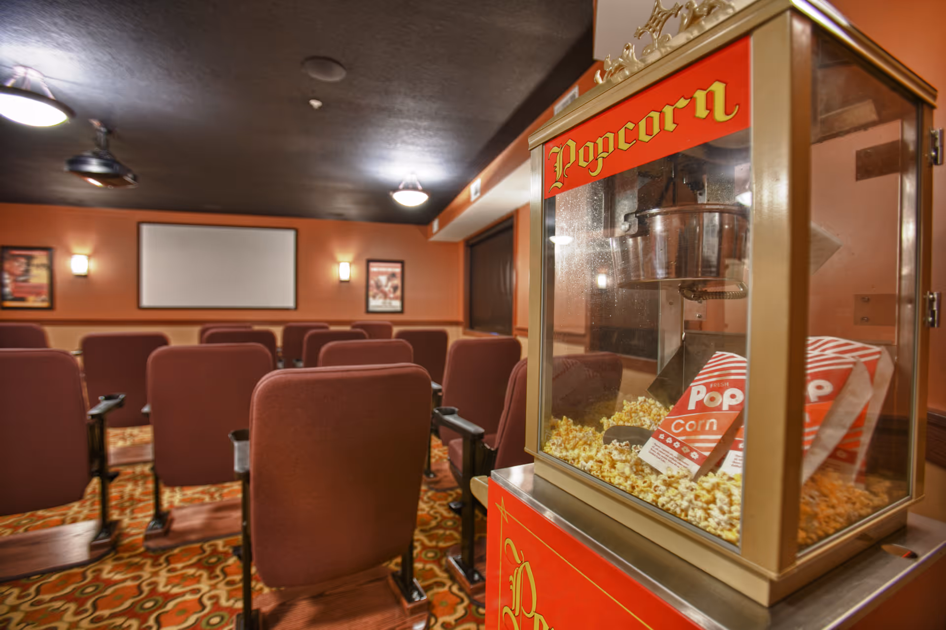 Small private movie theater with rows of upholstered seats, a popcorn machine in the foreground and a projection screen at the front.