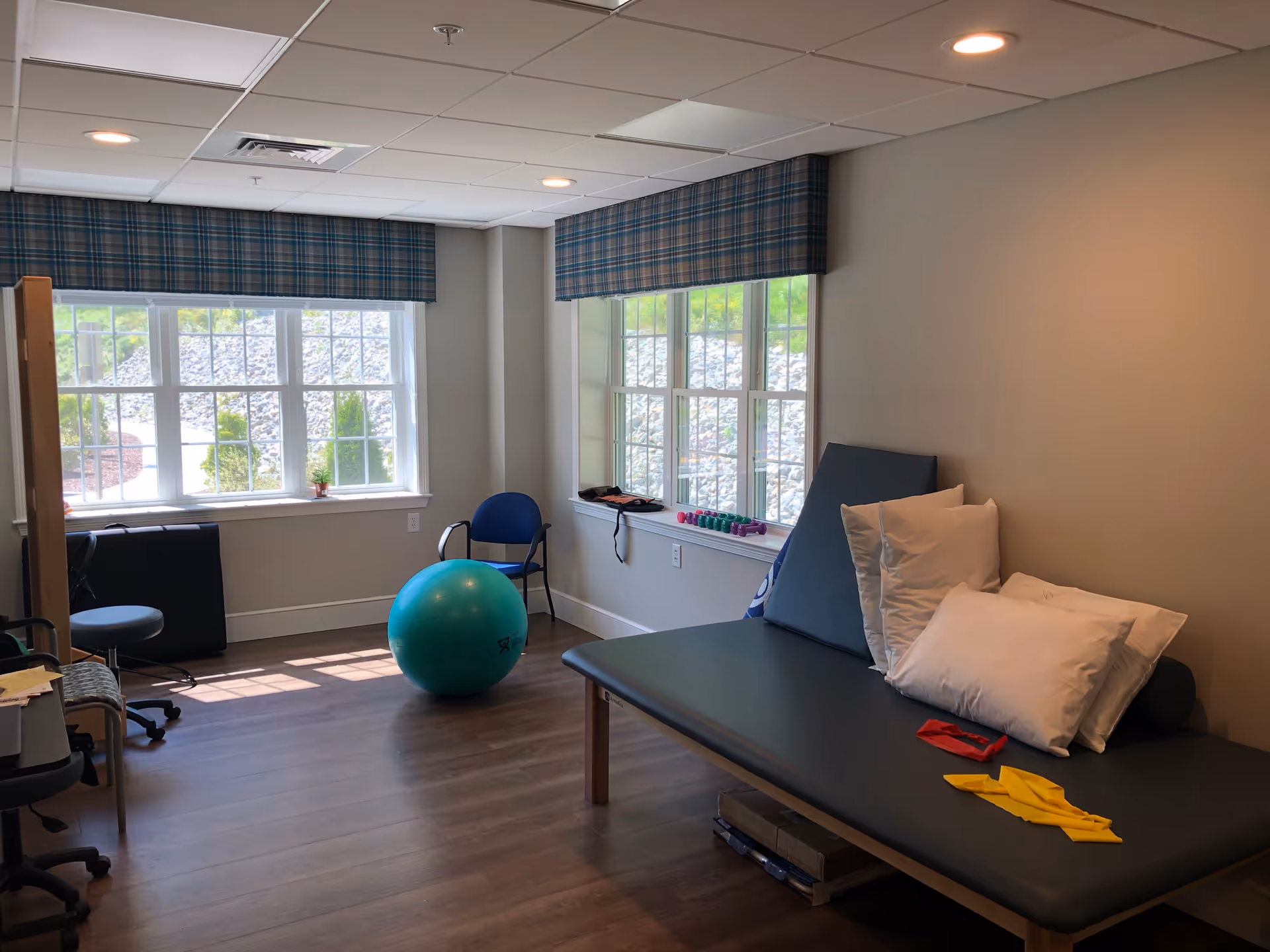 A therapy or exercise room with large windows letting in natural light. The room contains a blue exercise ball, a padded therapy table with several pillows, resistance bands, small dumbbells on the windowsill, and a few chairs. The floor is wood, and the walls are painted light beige.