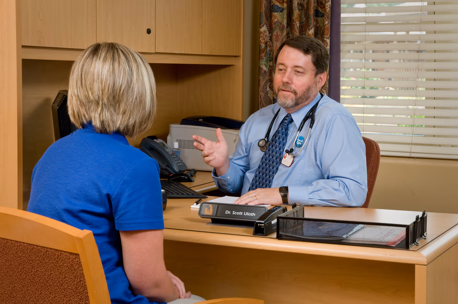 A male doctor with a stethoscope around his neck is sitting at a desk talking to a woman in a blue shirt. The doctor is gesturing with his hand while the woman listens attentively. The office has wooden furniture, a window with blinds, and a patterned curtain.
