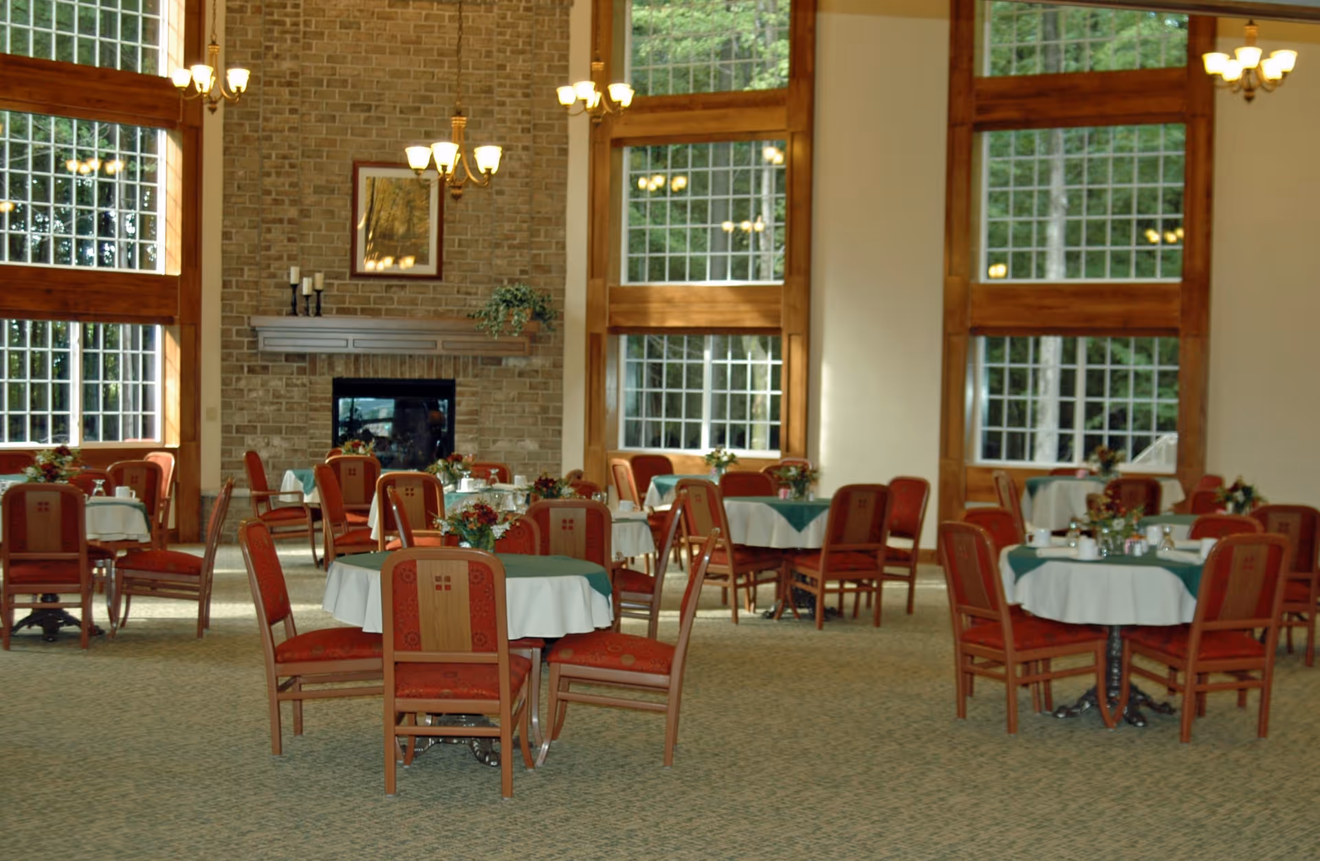 A spacious dining room with multiple round tables covered with white and green tablecloths, surrounded by red cushioned wooden chairs. The room features large windows with wooden frames allowing natural light to enter, a brick fireplace with a mantle decorated with candles and a plant, and chandeliers hanging from the ceiling.