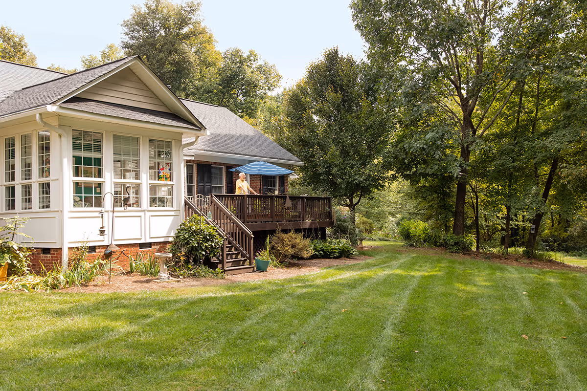 A sunny outdoor view of a senior living facility named Aldersgate showing a well-maintained lawn, trees, and a house with a porch where a person is standing under a blue umbrella.