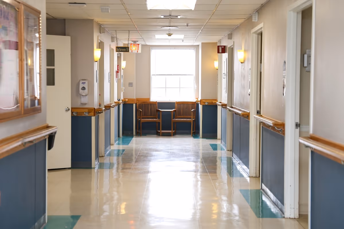 A clean, well-lit hallway in a senior living facility with beige and blue walls, handrails on both sides, several closed doors, two wooden chairs and a small table at the end near a large window, and an exit sign above the hallway.