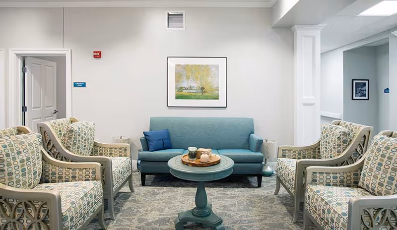 A cozy sitting area in a senior living facility with a teal sofa, four patterned armchairs, a round teal coffee table with candles, a framed landscape painting on the wall, and a light gray patterned carpet.