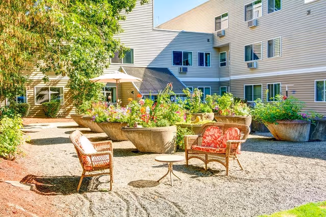 Outdoor courtyard area at Junction City Retirement and Assisted Living with large concrete planters filled with colorful flowers, wicker chairs with red floral cushions, a small round table, and a multi-story beige building in the background.