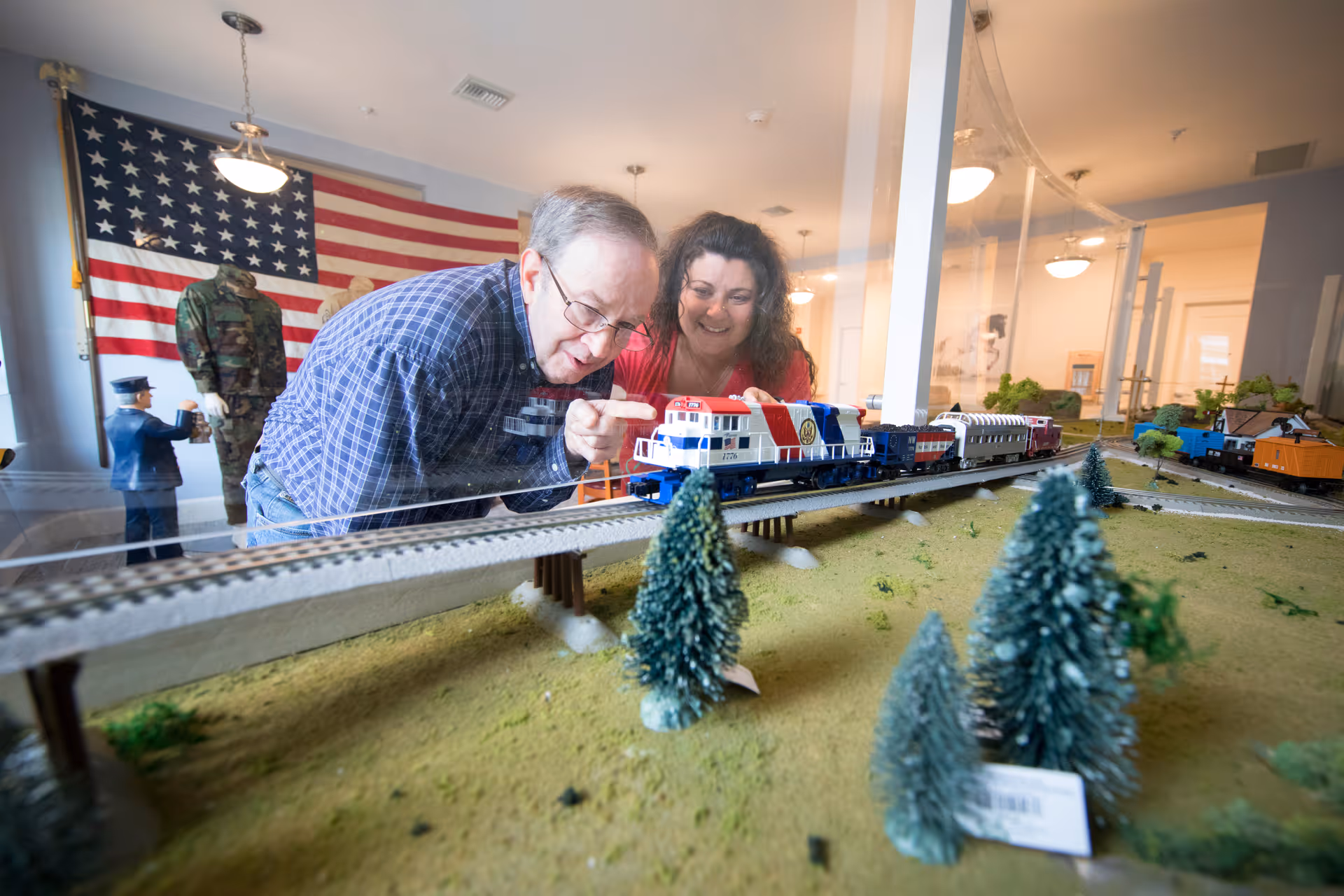 An elderly man and a woman are looking closely and smiling at a model train set displayed on a table. The scene is indoors with an American flag and military uniforms in the background.