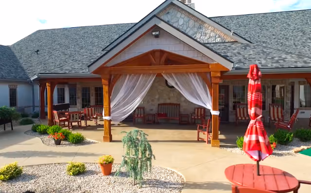 A paved courtyard with a wooden pergola draped in sheer curtains, red seating and umbrellas in front of a single-story building.