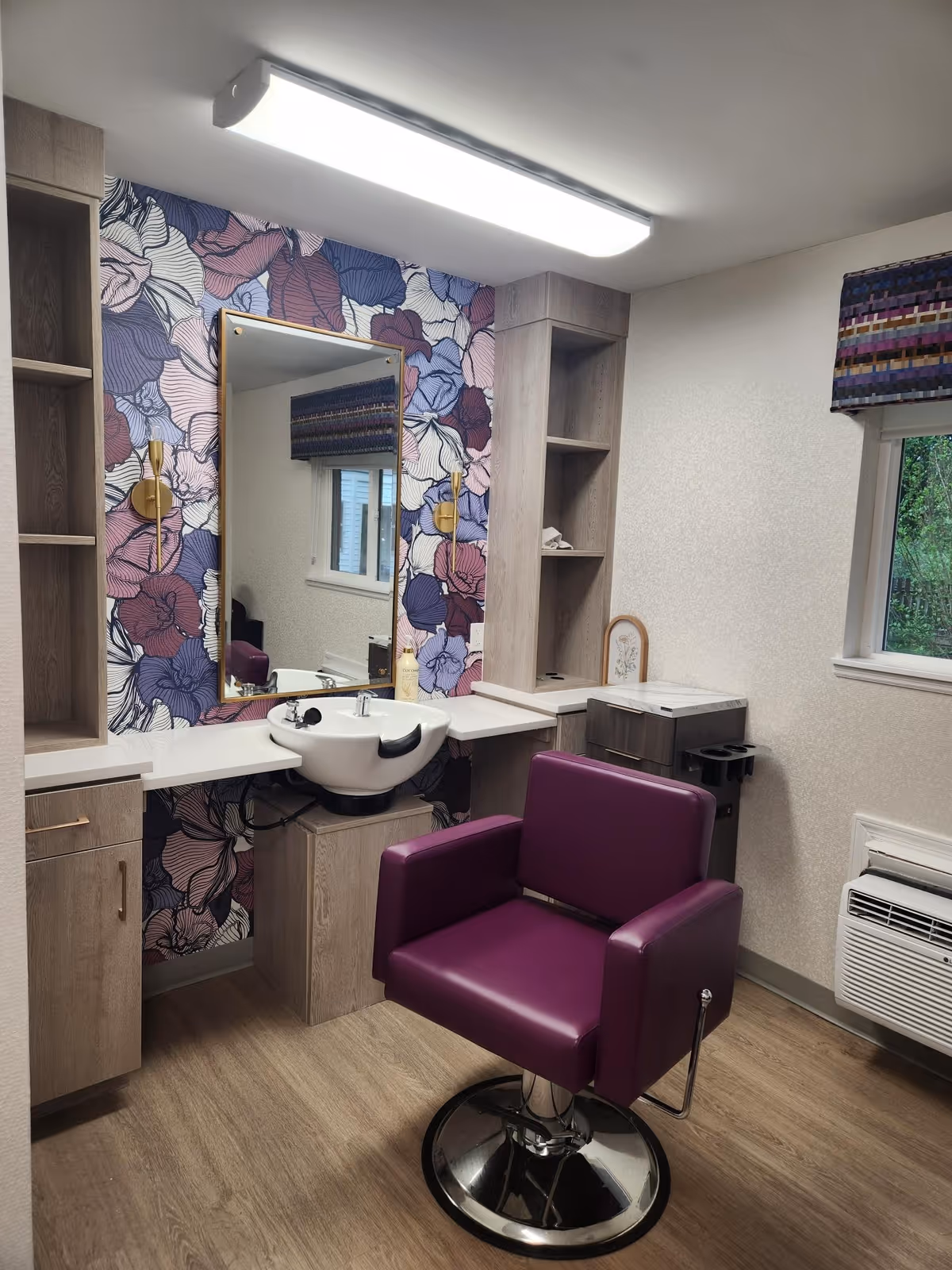 Interior view of a salon area in a healthcare facility featuring a purple salon chair in front of a white sink with a black neck rest. The wall behind the sink has colorful floral wallpaper with a large rectangular mirror and two gold wall sconces. There are wooden cabinets and shelves on either side of the sink, a window with a patterned valance, and a wall-mounted air conditioning unit below the window.