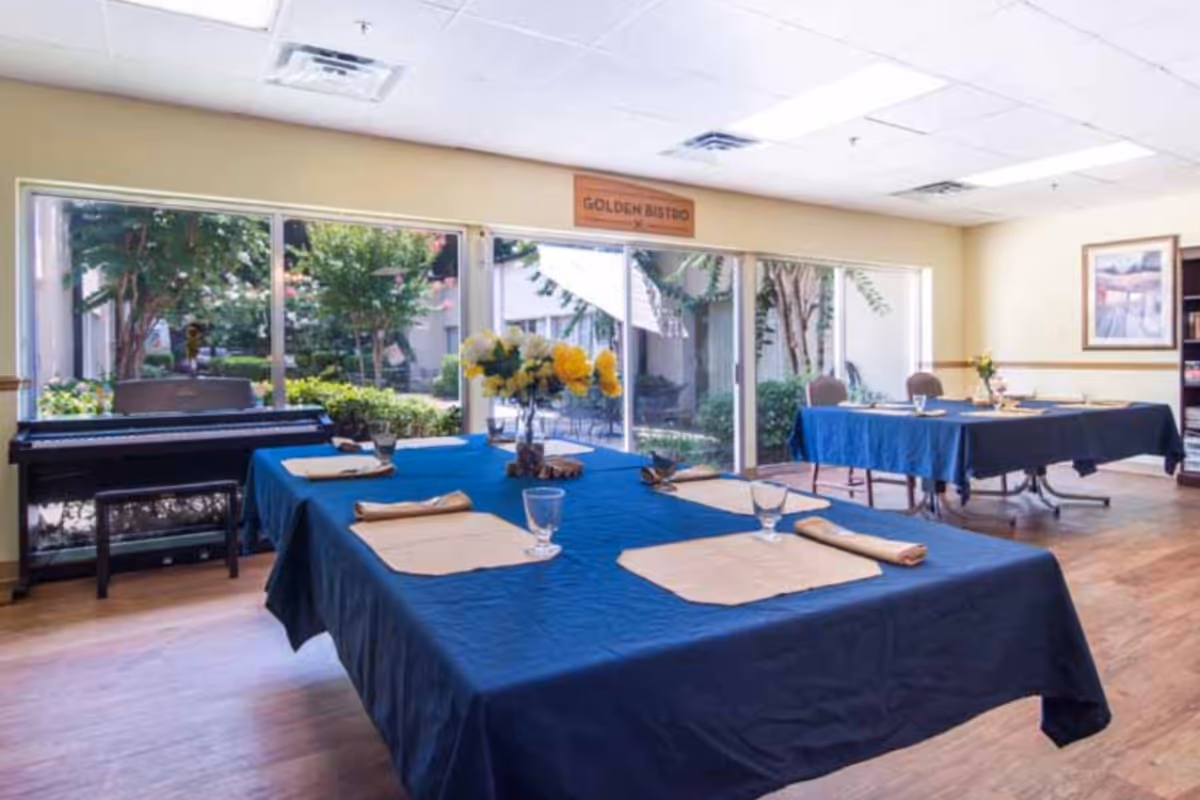 Dining room with two tables covered in blue tablecloths, each set with placemats, napkins, and glasses. A vase with yellow flowers is on the nearest table. Large windows show a garden outside, and a piano is positioned against the left wall. A sign above the windows reads 'GOLDEN BISTRO'.