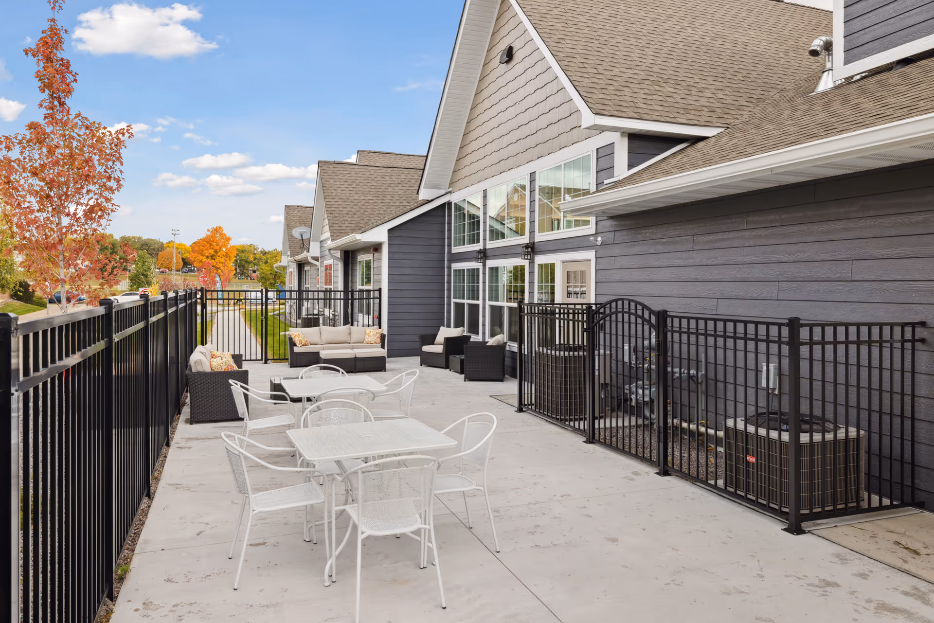 Outdoor patio area at a senior living facility with white metal tables and chairs, black cushioned seating, and a black metal fence. The building has large windows and gray siding, with autumn trees and a blue sky in the background.
