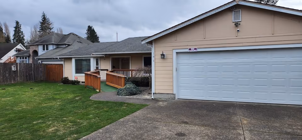 Exterior view of a single-story residential building with beige siding and a gray shingled roof. The building has a large white garage door on the right and a wooden ramp leading to the front door. The lawn is green and well-maintained, with a wooden fence on the left side and neighboring houses visible in the background under a cloudy sky.