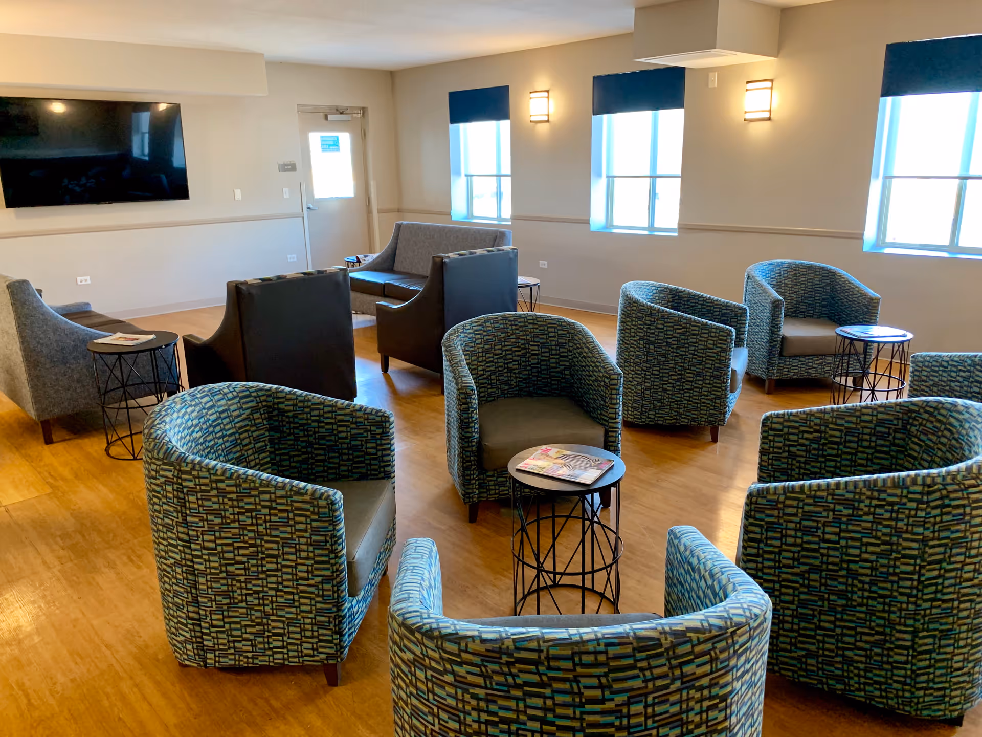 A spacious, well-lit lounge area with multiple patterned armchairs arranged around small round tables. There are also two gray sofas and two dark armchairs facing a large flat-screen TV mounted on the wall. The room has wooden flooring, three large windows with blue shades, and wall-mounted lights.