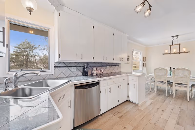 Bright kitchen with white cabinets, a stainless steel double sink, and a dishwasher. The countertop is gray with a tiled backsplash. A window above the sink shows trees outside. In the background, there is a dining area with a white dining table and chairs, and a modern chandelier hanging from the ceiling.