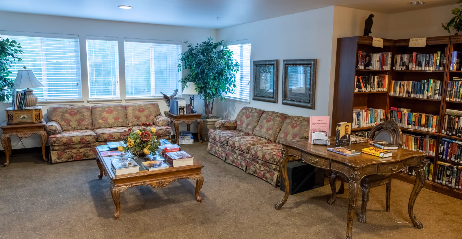 A cozy library or reading room with two floral patterned sofas, a wooden coffee table with books and a flower arrangement, a wooden desk with books and papers, and bookshelves filled with books. The room has large windows with blinds, two framed pictures on the wall, and potted plants adding greenery.