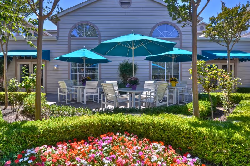 Outdoor patio area with several white tables and chairs under teal umbrellas, surrounded by green shrubs, trees, and colorful flowers in front of a beige building with large windows and teal awnings.
