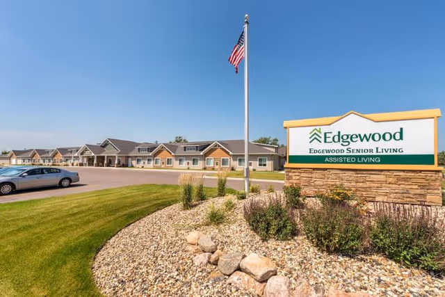 Exterior view of Edgewood Senior Living assisted living facility with a large sign in the foreground, an American flag on a flagpole, a parking lot with a car, and a row of single-story buildings under a clear blue sky.