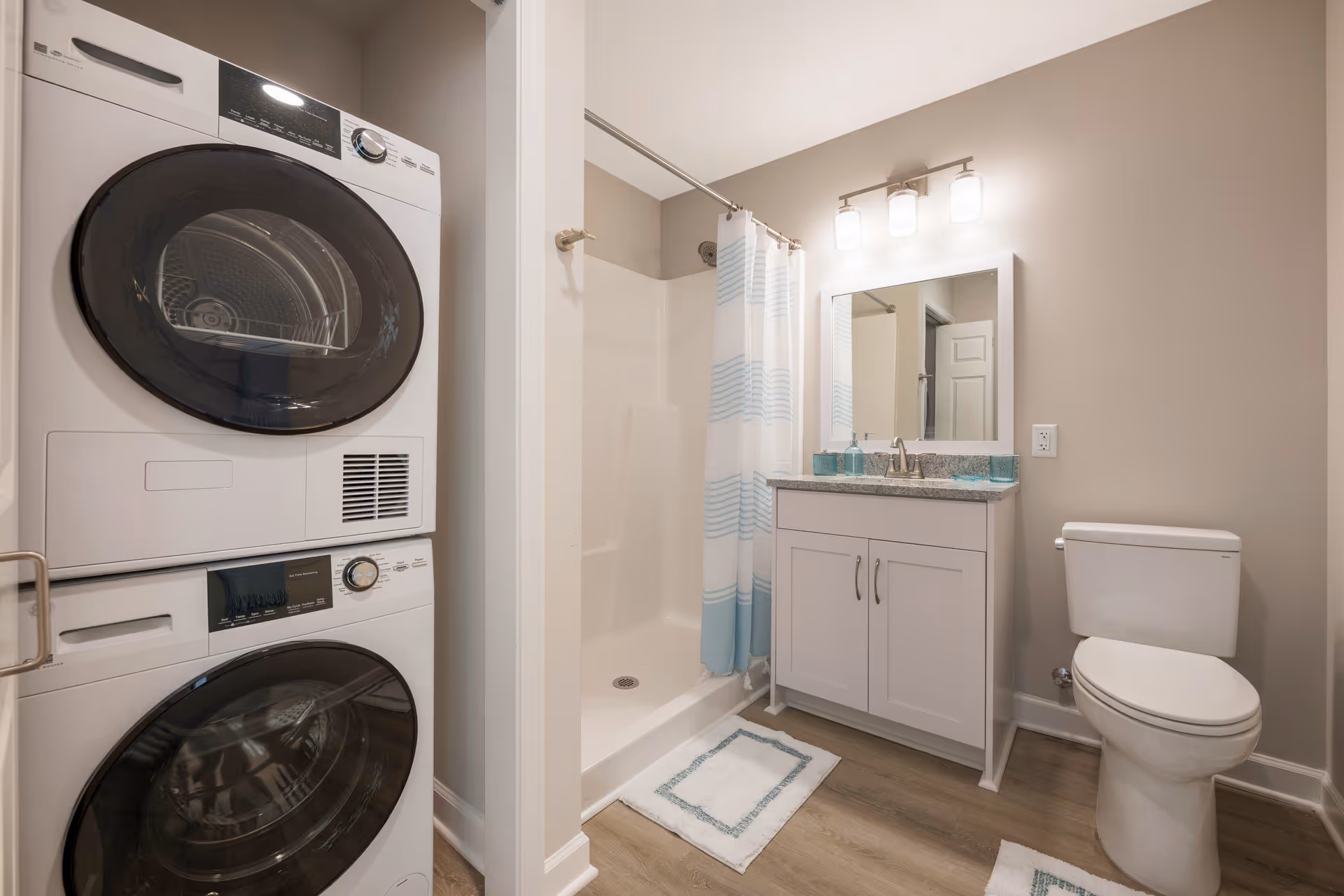 A bathroom featuring a white toilet, a vanity with a sink and mirror above it, a shower with a blue and white striped curtain, and a stacked washer and dryer unit to the left. The room has light-colored walls and wood-style flooring.