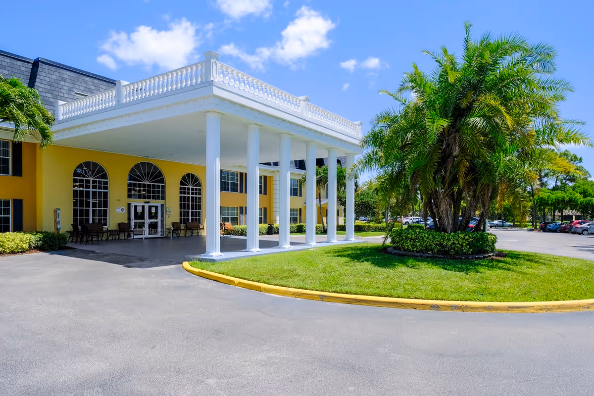 Front exterior view of Volante Senior Living of Delray Beach showing a yellow building with large white columns supporting a covered entrance. There are palm trees and green landscaping in front, with a parking lot visible to the right under a blue sky with some clouds.