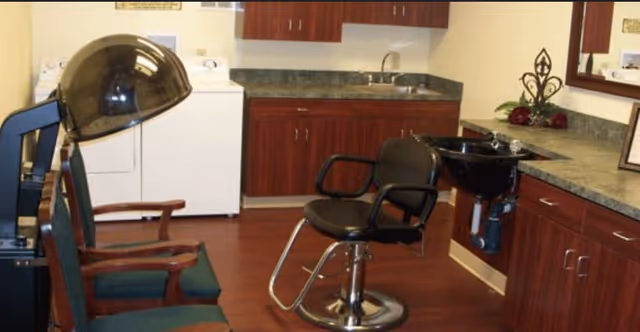 Interior view of a salon area within a senior living facility featuring a black salon chair in front of a black wash basin, two green cushioned chairs with wooden armrests, a hair dryer hood, and wooden cabinets with a countertop and sink in the background.