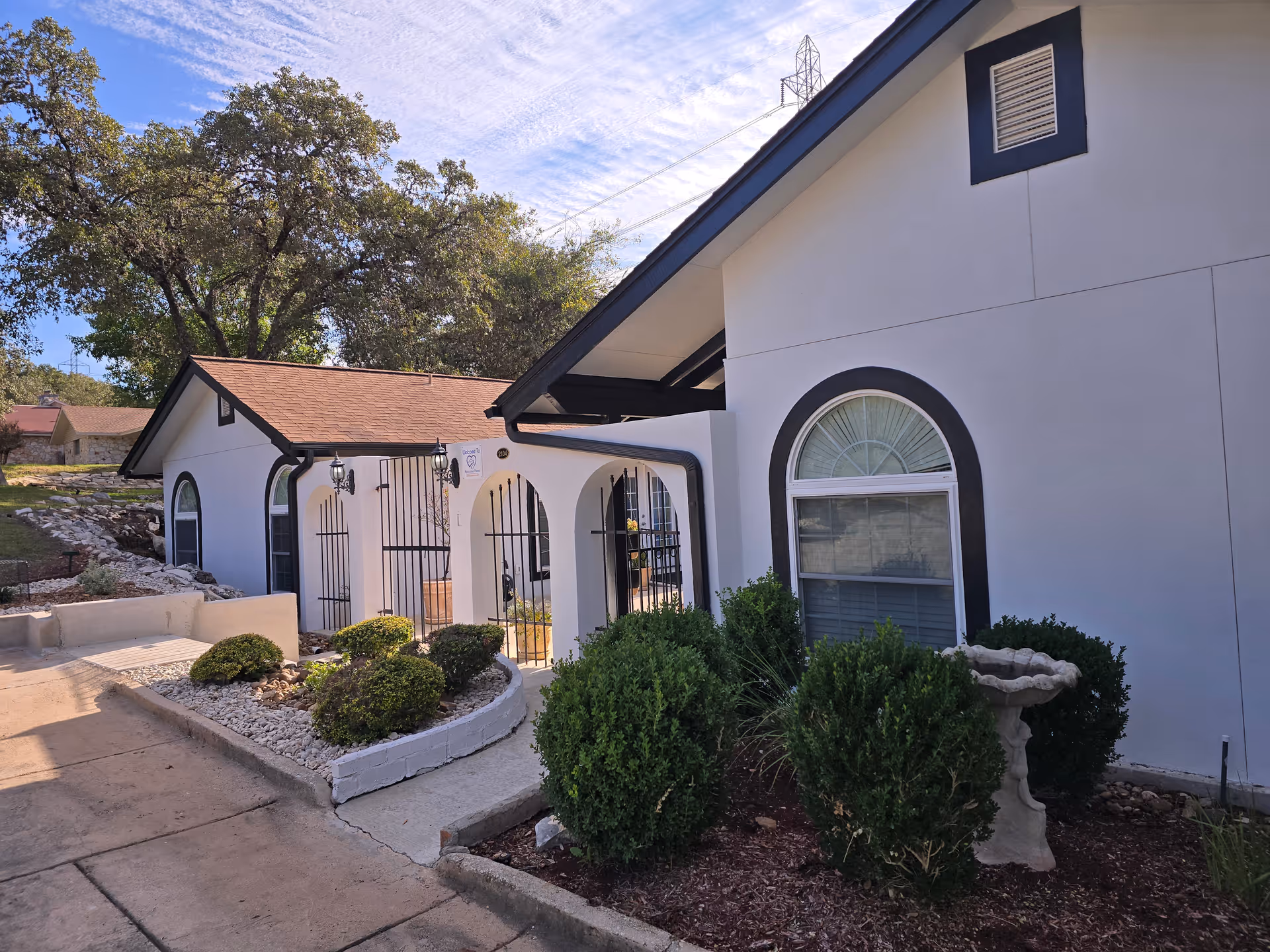 Exterior front of a white single-story building with arched windows, a gated arched porch, and landscaped shrubs and a walkway.