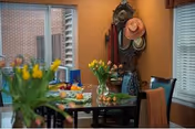 A cozy dining area with a glass table set with plates, fruit, and a vase of yellow tulips. In the background, there is a wooden hall tree with hats and a mirror, and windows with blinds letting in natural light.