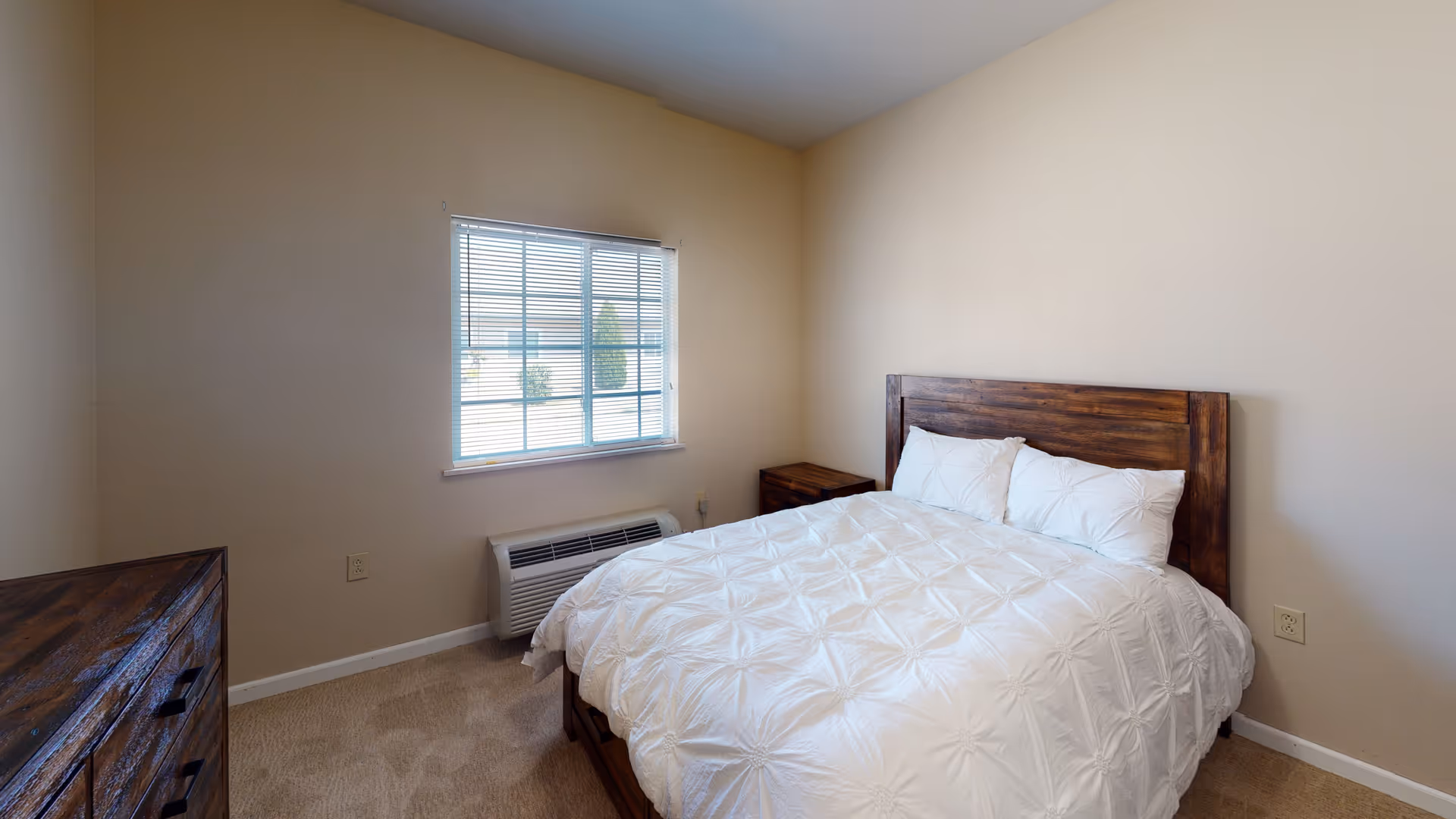A simple bedroom with beige walls and carpeted floor. The room features a wooden bed frame with a white comforter and three pillows. There is a wooden nightstand beside the bed and a wooden dresser on the left side. A window with white blinds lets in natural light, and an air conditioning unit is installed below the window.