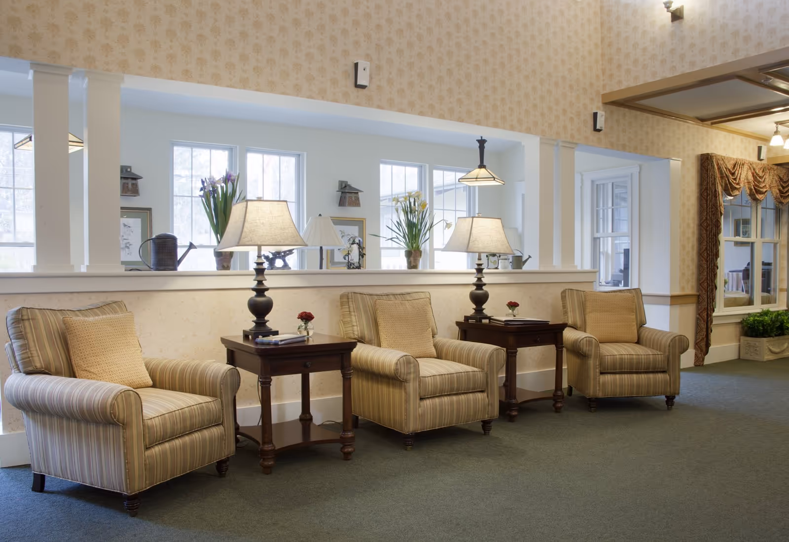 Three upholstered armchairs with side tables and lamps arranged in a bright, carpeted sitting area with windows and decorative plants.