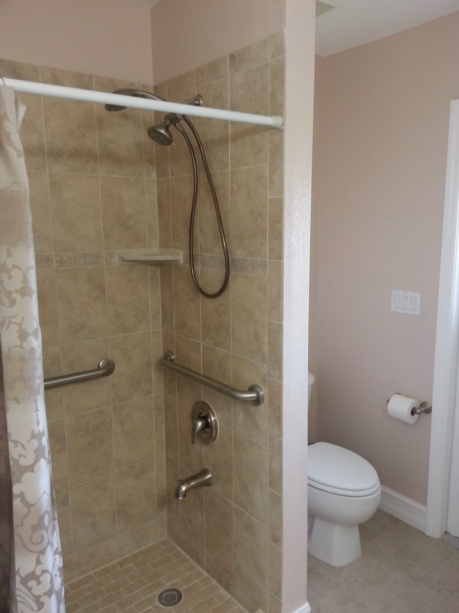 A bathroom with a tiled walk-in shower featuring two grab bars, a handheld showerhead, and a shower curtain. Next to the shower is a toilet with a toilet paper holder mounted on the wall.