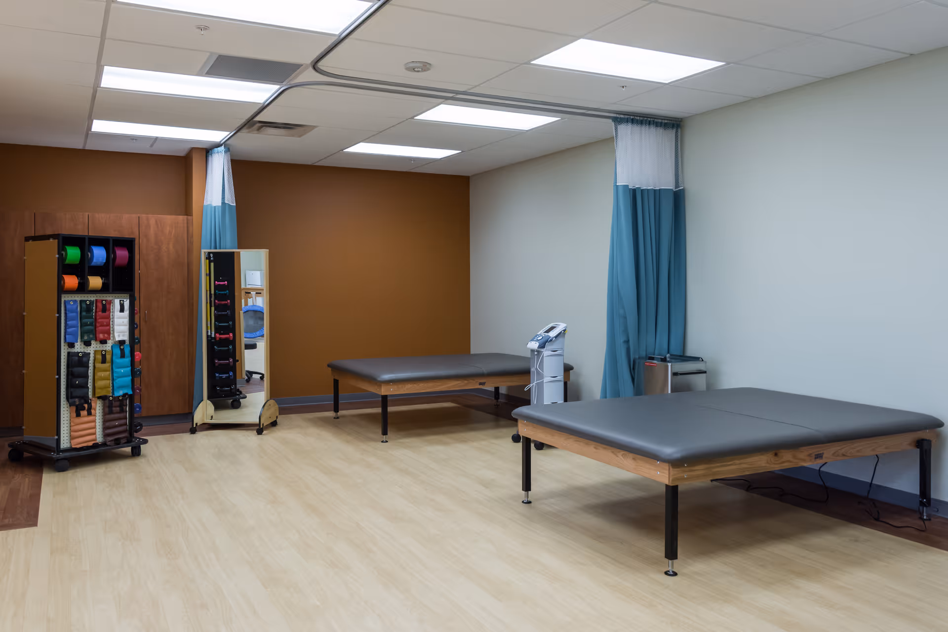 A rehabilitation therapy room with two padded treatment tables, equipment storage racks, and privacy curtains.