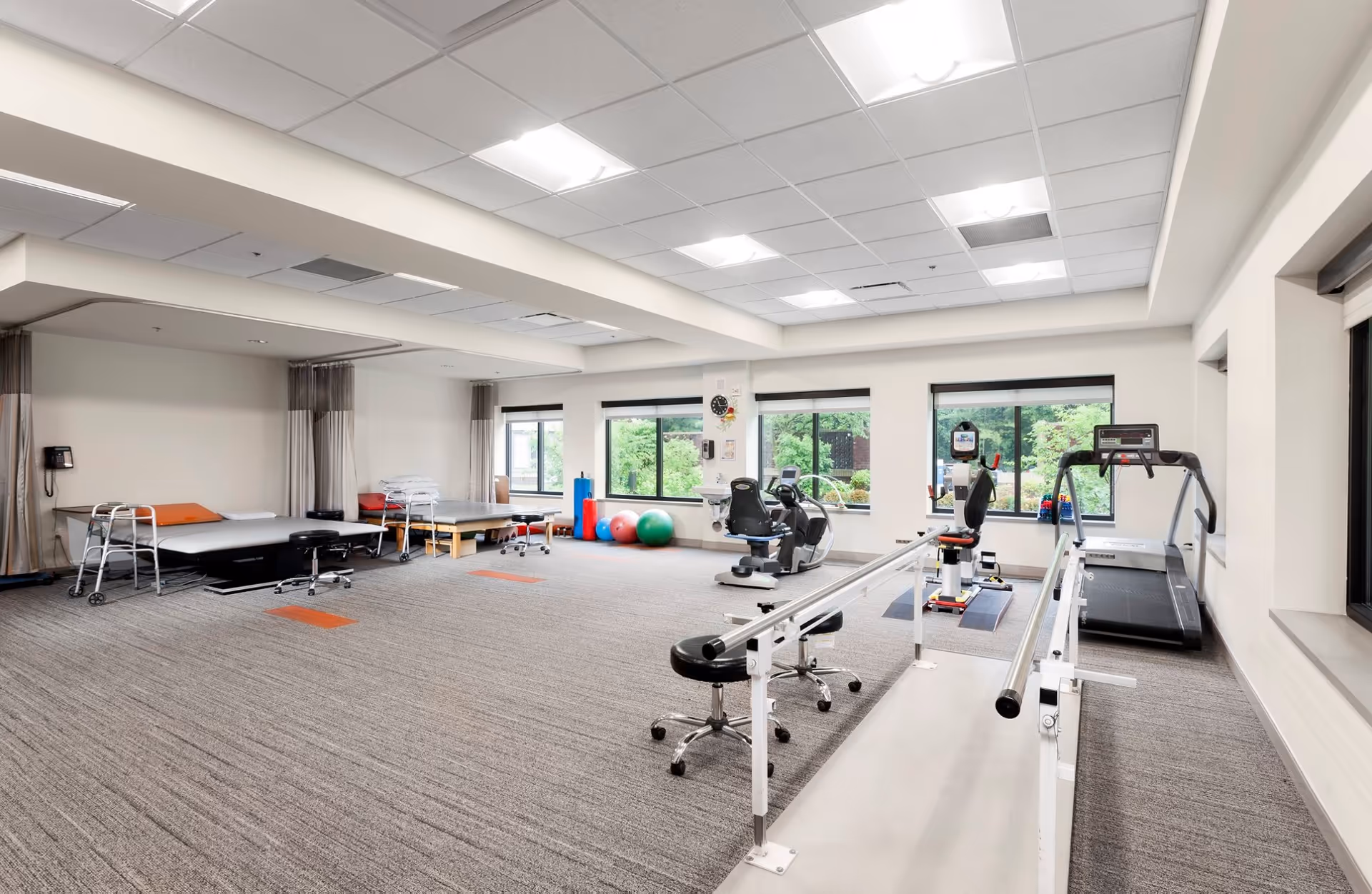 A spacious physical therapy room with exercise equipment including a treadmill, stationary bikes, parallel bars for walking practice, therapy beds with walkers, exercise balls, and stools. Large windows provide natural light and a view of greenery outside.