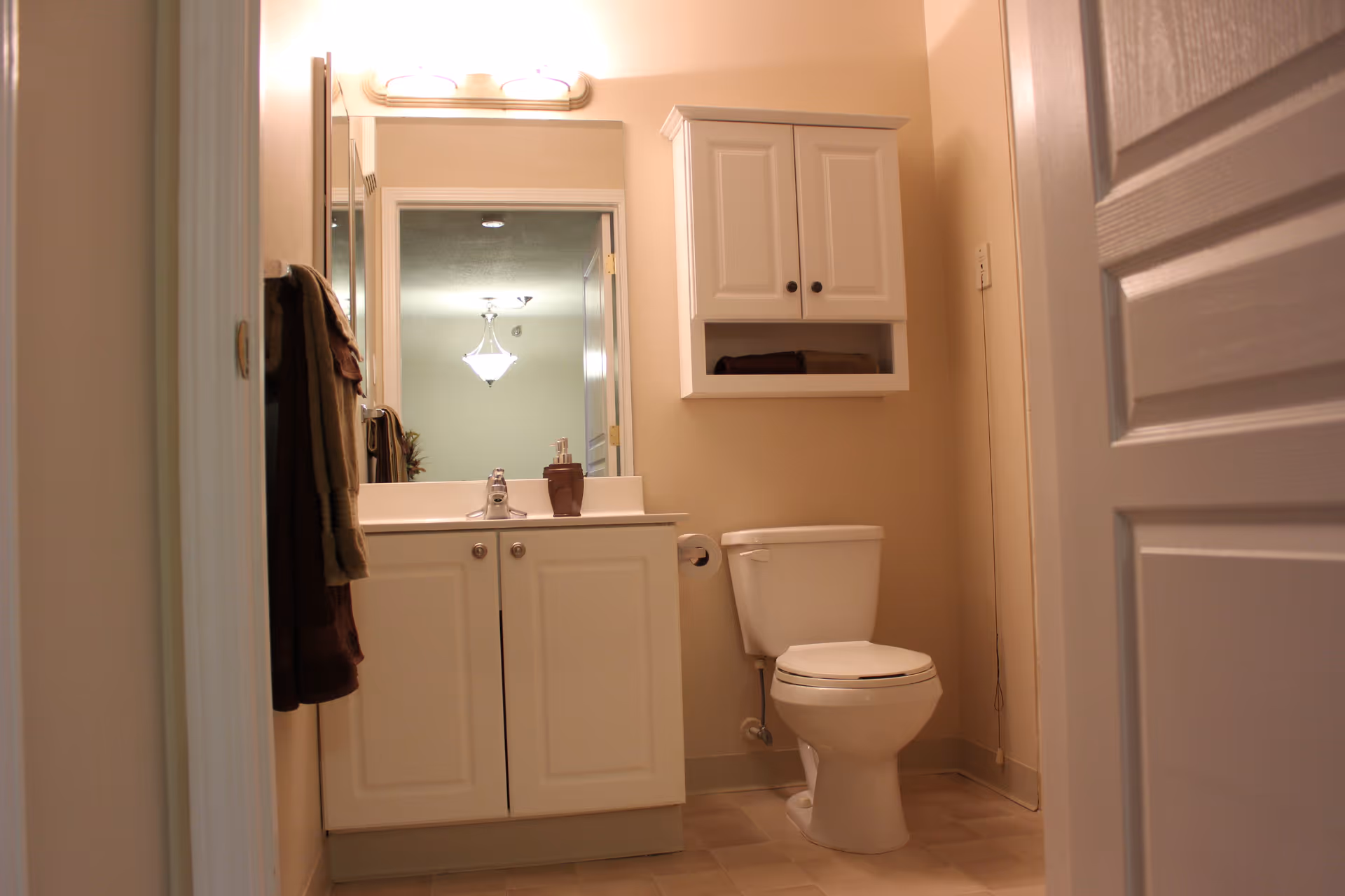 Bathroom with a white sink vanity and mirror, wall-mounted cabinet, toilet, and towels hanging by the door.