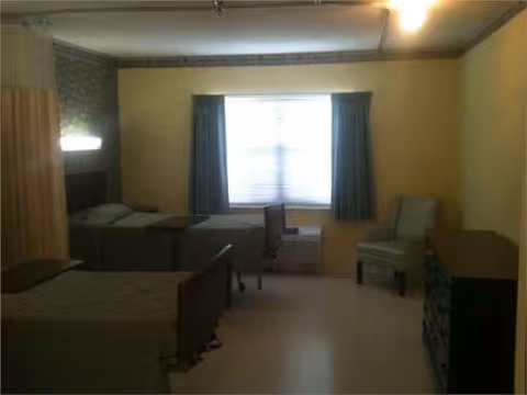 Two beds and seating in a small dim bedroom with a window and blue curtains.