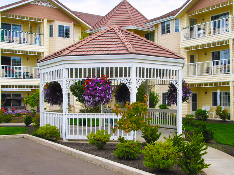 A white wooden gazebo with a brown shingled roof surrounded by green shrubs and hanging baskets of purple and red flowers, situated in front of a yellow multi-story residential building with balconies.
