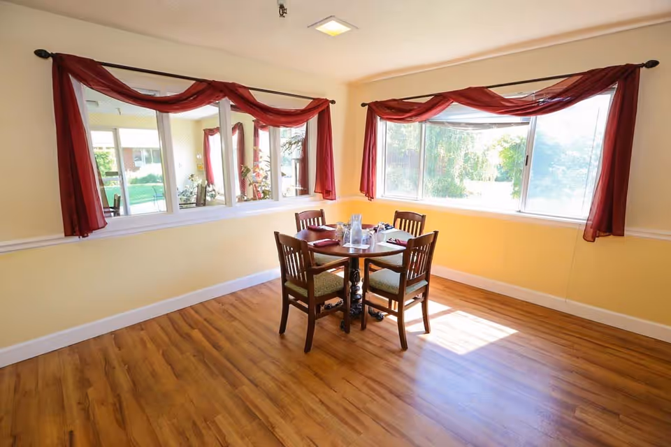 A small dining area with a round wooden table set for four with napkins and a water pitcher, surrounded by four wooden chairs with green cushions. The room has light yellow walls, large windows with red curtains, and wood flooring. Sunlight streams in through the windows, creating a bright and inviting atmosphere.