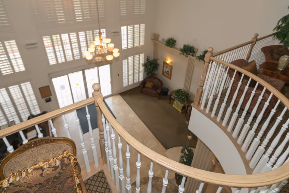 View from an upper level overlooking a senior living facility's lobby area with a curved wooden and white railing, a chandelier, large windows with shutters, a leather armchair, plants, and framed artwork on the walls.