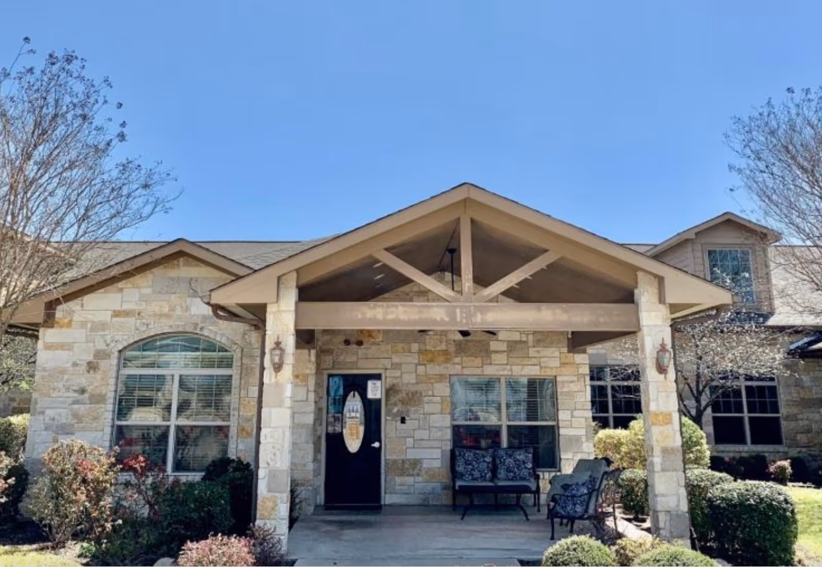 Front exterior view of a stone building with a covered entrance supported by two pillars. There are two windows on either side of a black door with an oval glass panel. Outdoor seating with cushioned chairs is placed under the covered entrance. The building is surrounded by neatly trimmed bushes and trees under a clear blue sky.