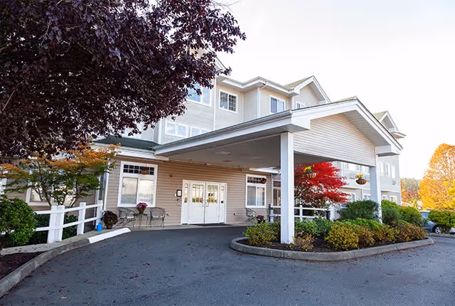 Exterior view of Artesian Place Assisted Living facility showing the main entrance with a covered driveway, surrounded by landscaped bushes and trees with autumn foliage.