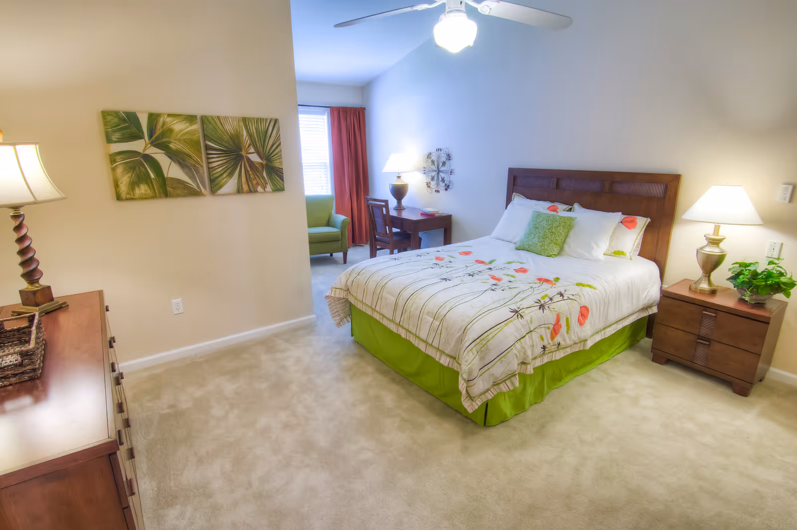 A bright and cozy bedroom with a bed featuring floral bedding and a green bed skirt. The room includes two wooden nightstands with lamps, a wooden dresser with a lamp and basket, a small desk with a chair and lamp, a green armchair near a window with red curtains, and two framed leaf paintings on the wall.