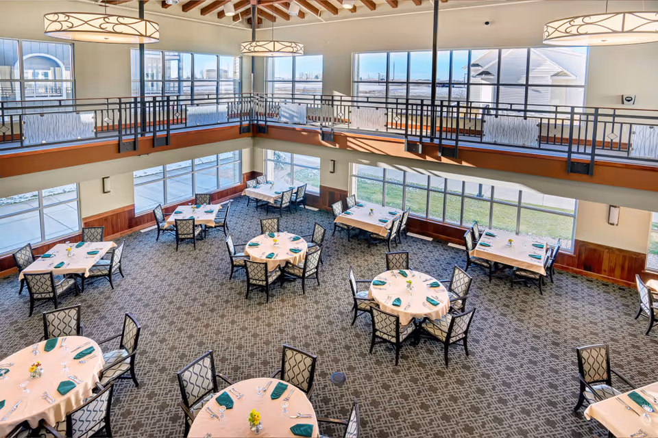 A spacious, well-lit dining room with multiple round and rectangular tables covered with beige tablecloths, each set with green napkins, silverware, and small floral centerpieces. Large windows surround the room, allowing natural light to fill the space, and a second-floor balcony overlooks the area. Modern pendant lights hang from the ceiling.