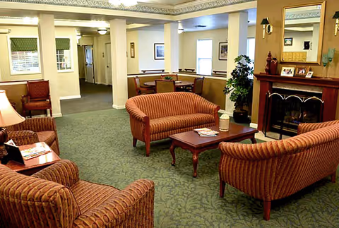 A cozy living room area in a senior living facility with striped upholstered armchairs and sofas arranged around a wooden coffee table. There is a fireplace with a mirror above it on the right side, a potted plant nearby, and a dining area visible in the background. The room has green patterned carpet and soft lighting.