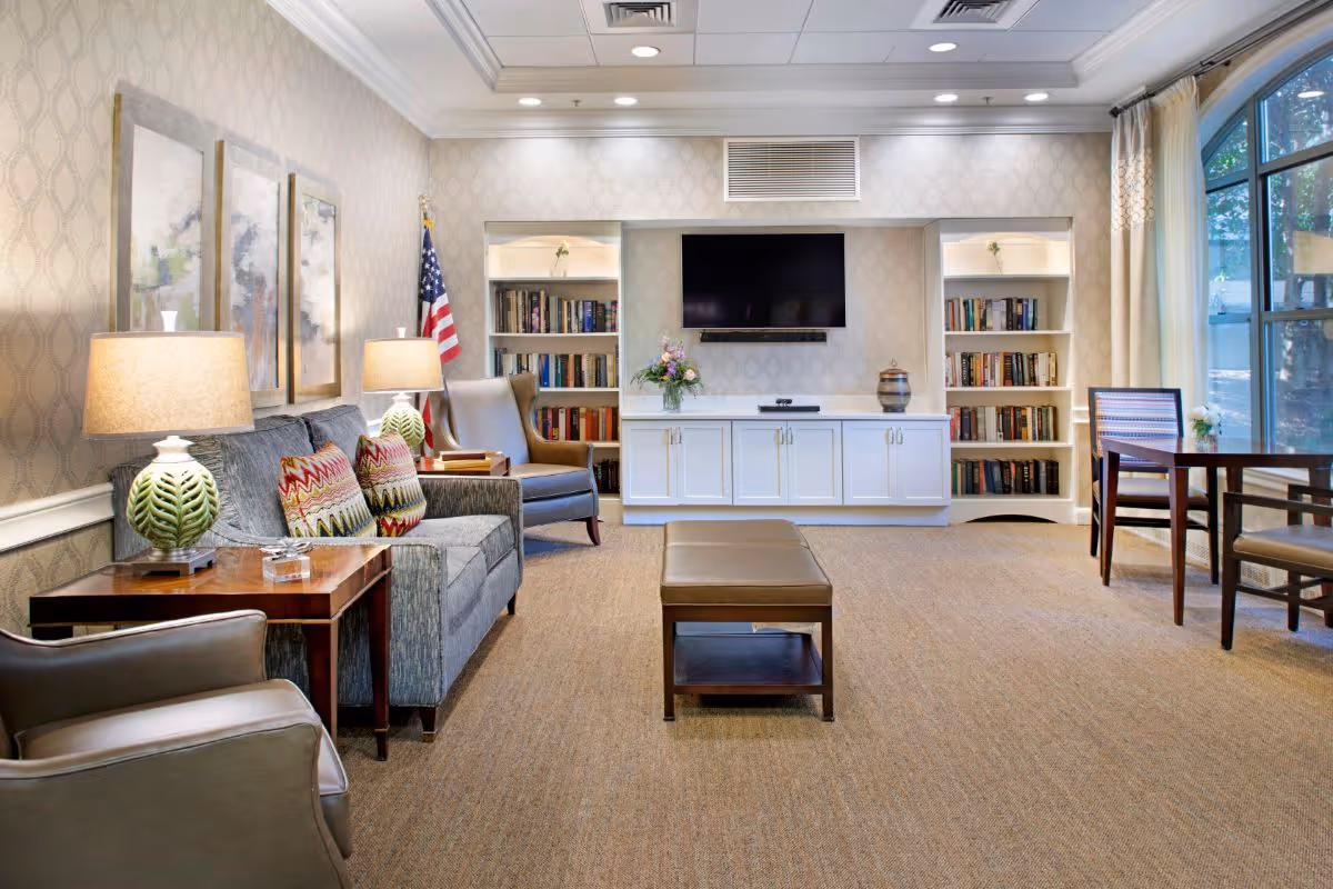 A cozy living room with a gray sofa adorned with colorful patterned pillows, two armchairs, and a wooden coffee table. There are two table lamps with green bases on side tables next to the sofa. The back wall features built-in white cabinets and bookshelves filled with books, a flat-screen TV mounted above the cabinets, and a vase with flowers. An American flag stands in the corner, and large windows with curtains allow natural light to brighten the room.