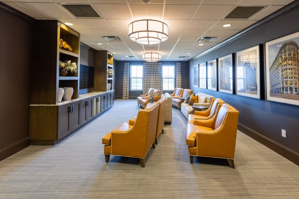 A spacious, well-lit lounge area with multiple mustard yellow armchairs and beige sofas arranged in two rows facing each other. The room features a long wooden built-in cabinet with decorative items and a flat-screen TV mounted on the wall. Large windows with patterned curtains allow natural light to fill the room. The walls are adorned with framed architectural photographs, and two modern ceiling light fixtures hang overhead.