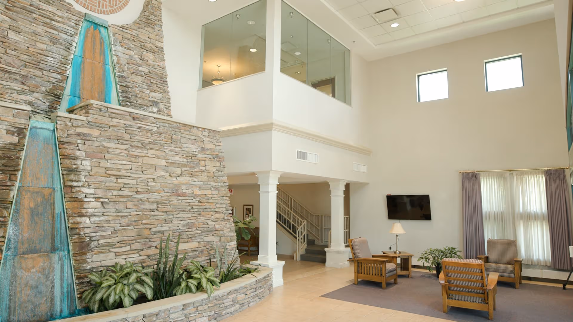 A spacious living room area with high ceilings and large windows covered by light curtains. The room features a stone wall with a decorative water feature and plants at its base. There are wooden chairs with cushions arranged around a small table with a lamp, and a flat-screen TV mounted on the wall. A staircase with a railing leads to an upper floor with glass walls.