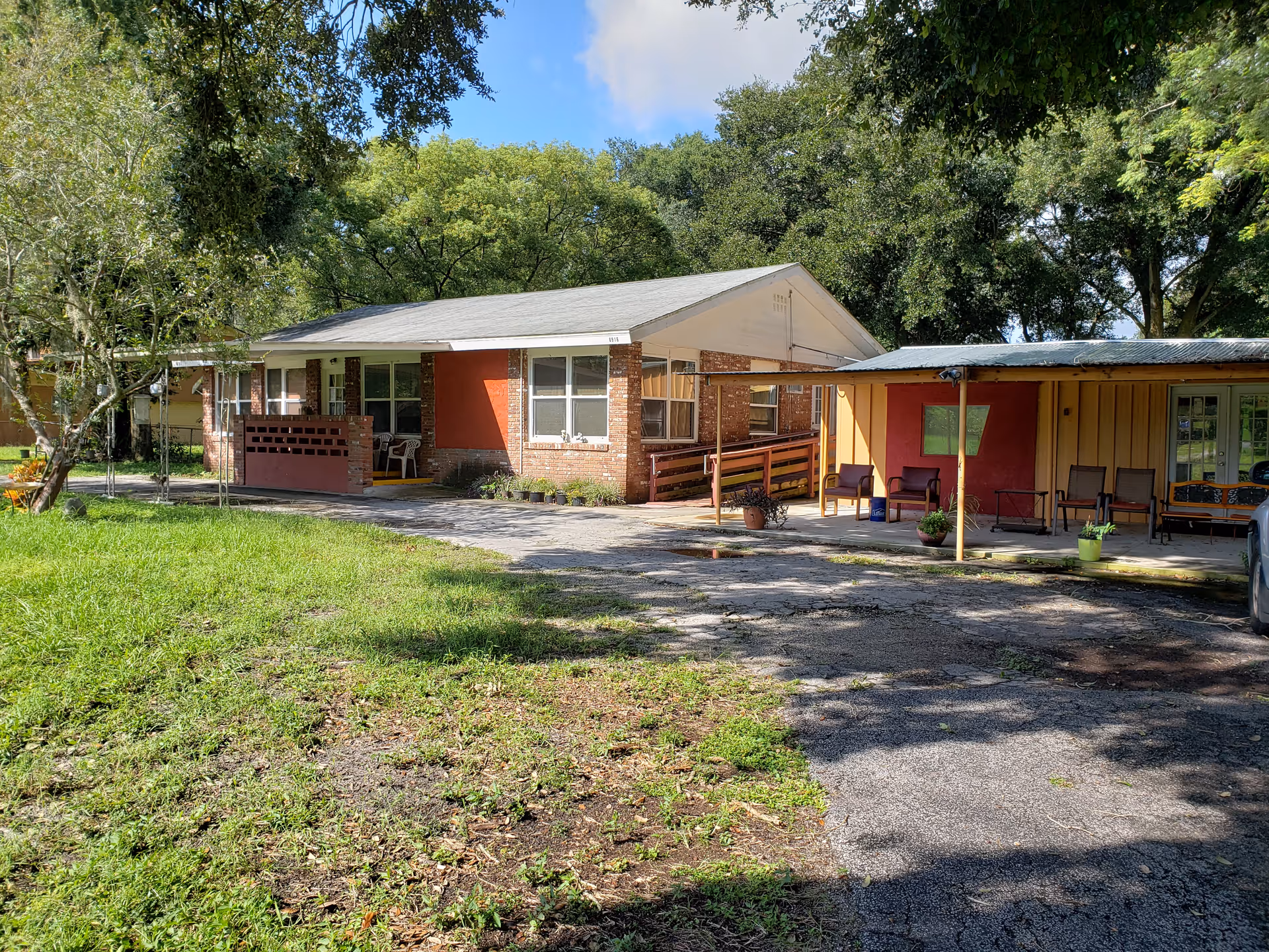 Single-story brick and wood building with a covered porch area furnished with chairs, surrounded by green grass and trees under a blue sky with some clouds.