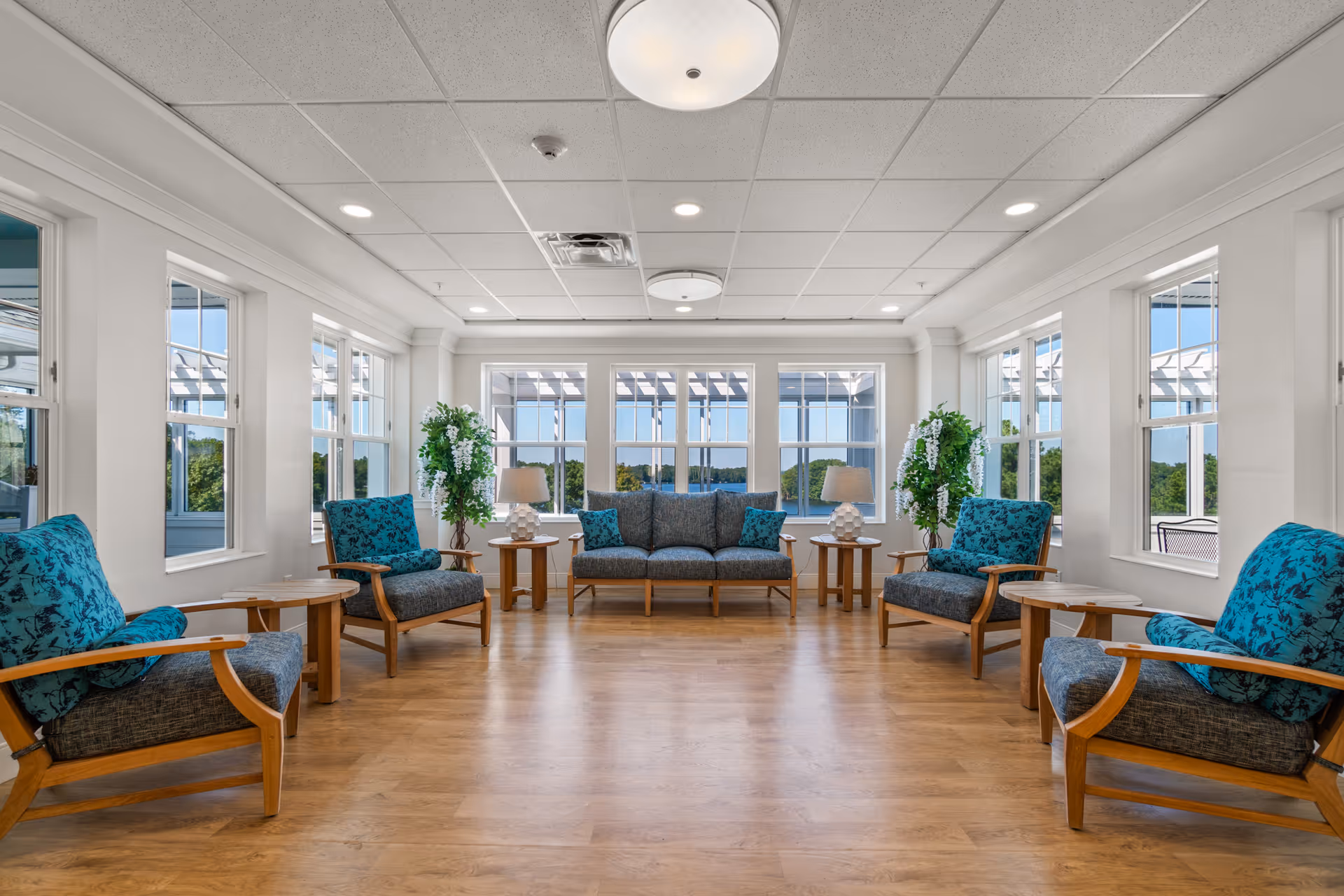 A bright and spacious living room with large windows on three sides letting in natural light. The room features a wooden floor, a central blue upholstered sofa flanked by two matching armchairs on each side, all with wooden frames and blue patterned cushions. There are small wooden side tables with lamps and green potted plants near the windows. Outside the windows, a pergola and greenery are visible.