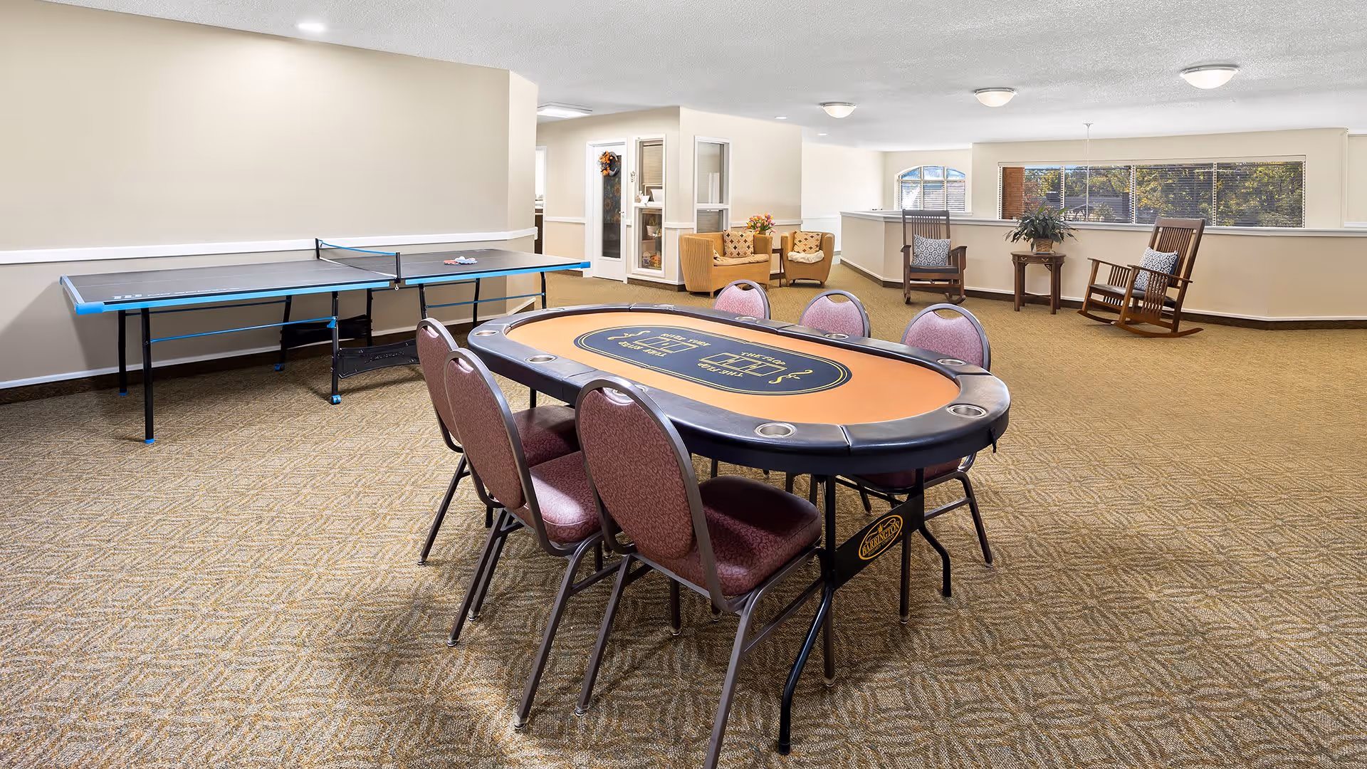 A spacious recreational room with a poker table surrounded by six chairs in the center. To the left, there is a ping pong table against the wall. In the background, there are two armchairs with a small table between them, and two rocking chairs near a large window with a view of trees outside. The room has beige walls and carpeted flooring with a geometric pattern.